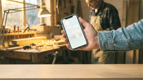Editorial lifestyle photo: a craftsperson's hands holding a phone in a workshop during an evening, warm window light, sc